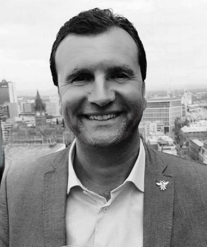 A man in a light gray suit smiles against a city skyline backdrop. The photo is in black and white, highlighting his cheerful expression.