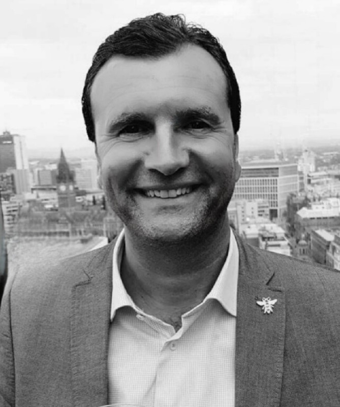 A man in a light gray suit smiles against a city skyline backdrop. The photo is in black and white, highlighting his cheerful expression.