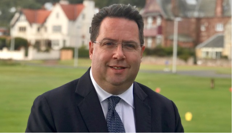 A man in a dark suit and glasses smiles confidently against a backdrop of a green lawn and elegant buildings.