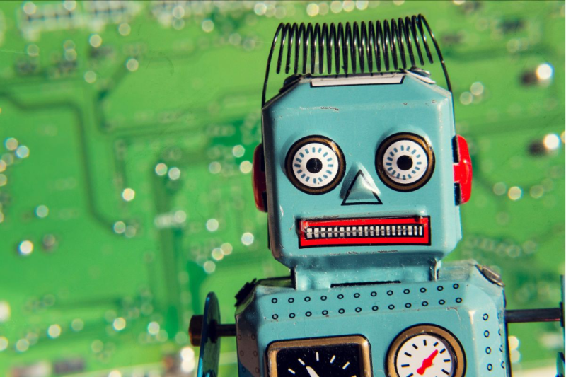 A vintage toy robot with a blue body, red ears, and a clock on its chest, set against a blurred green circuit board background. The robot has large expressive eyes and a unique hairline made of spring.