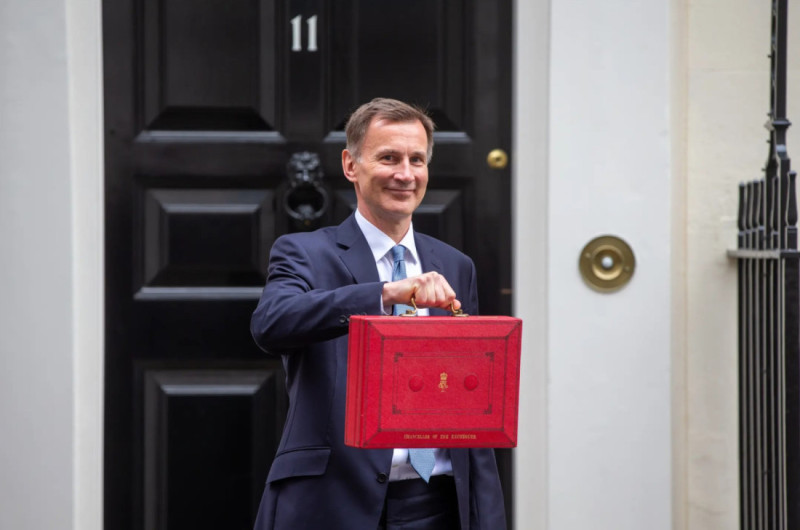 A man in a suit stands in front of a black door number 11, holding a red briefcase with a confident smile.