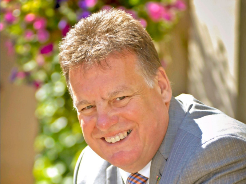 A man smiling warmly, wearing a gray suit and orange tie, with vibrant flowers in the background.