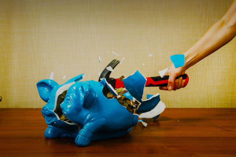 A blue piggy bank is being smashed open with a wrench, with coins spilling out and pieces of the piggy bank flying in various directions.