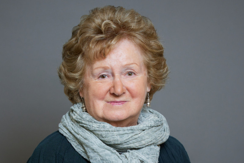 An elderly woman with curly, light brown hair smiles warmly, wearing a light gray scarf over a dark top, against a neutral gray background.