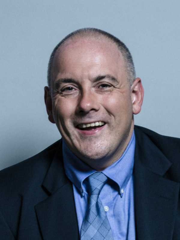 A smiling man with short, bald hair wearing a dark suit and a light blue tie against a plain backdrop.