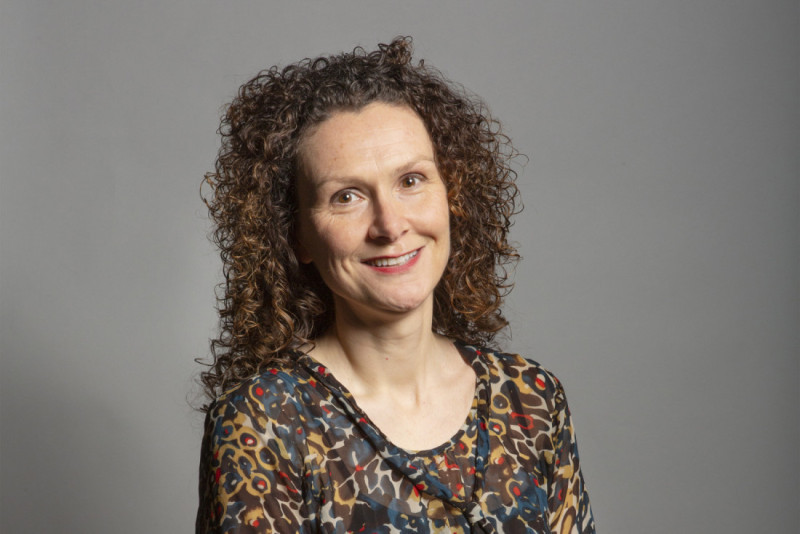 A woman with curly hair wearing a patterned blouse smiles warmly against a gray background.