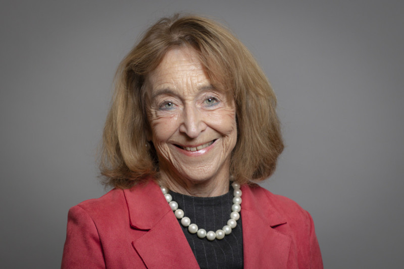 A smiling woman with shoulder-length hair, wearing a red blazer and a pearl necklace, poses against a gray background.