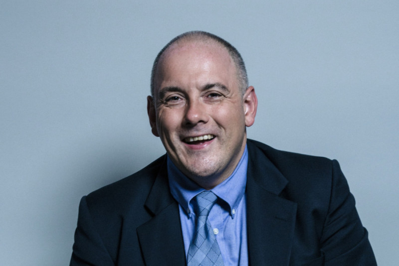 A smiling man with short bald hair, wearing a dark suit, light blue shirt, and a patterned tie, against a light gray background.