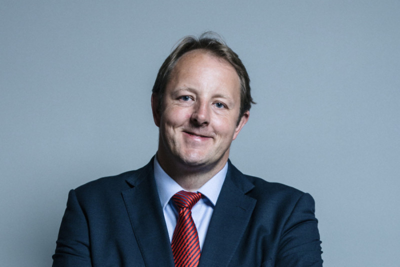 A man in a dark suit with a light blue shirt and red tie smiles confidently against a gray background.