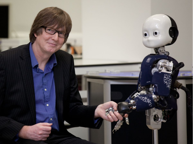 A researcher in a suit sits next to a humanoid robot, smiling as he shakes its hand. The robot has a small head and a torso with a blue and white design.