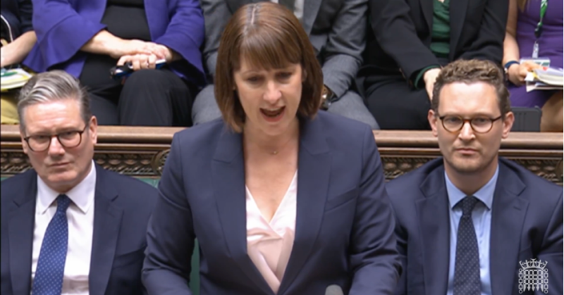 A woman in a dark blazer passionately addresses a parliamentary session, flanked by two men in suits. The background shows other attendees in formal attire, focusing on the speaker.