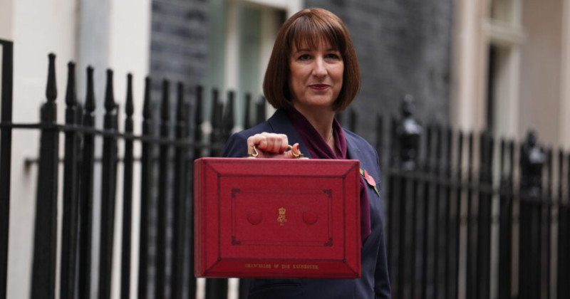 A woman with shoulder-length brown hair holds a red budget briefcase outside a government building, smiling confidently.