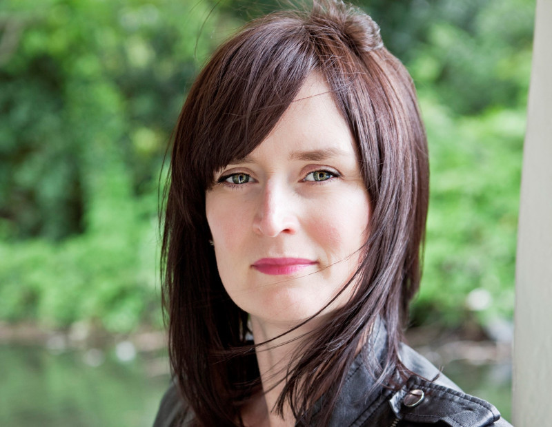 A woman with long brown hair and a leather jacket stands outdoors, facing the camera with a slight smile. Green foliage is blurred in the background.