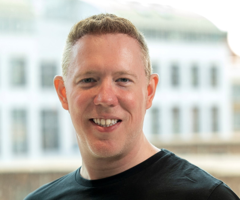 A smiling man with short, light brown hair wearing a black t-shirt, standing in front of a blurred background of a building.