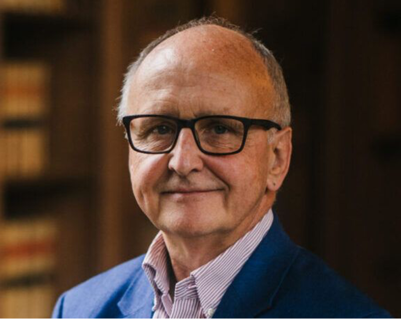 A middle-aged man with glasses and a friendly smile stands in front of blurred wooden shelves. He is wearing a blue blazer over a collared shirt.