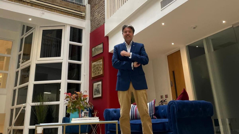 Rupert Pleasant, CEO of Guernsey Finance stands confidently in a modern, airy living space with large windows. He is dressed in a blue blazer and beige pants, surrounded by stylish furniture and decorative plants.