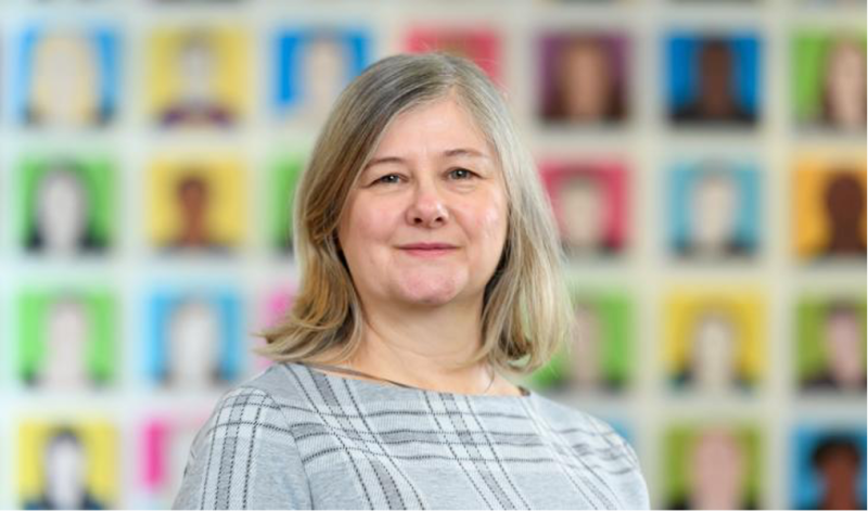 A woman with shoulder-length gray hair and a plaid gray top stands in front of a colorful wall featuring a grid of portraits.