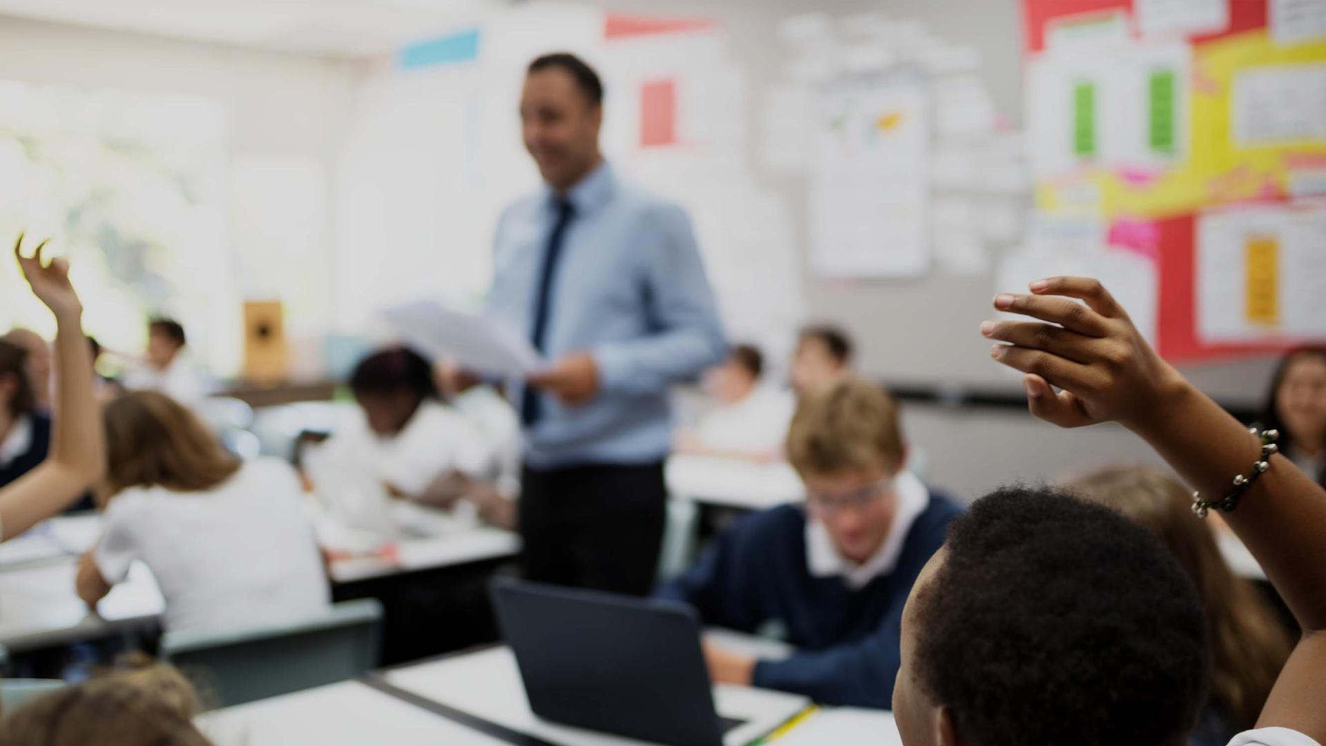 A classroom scene with students seated at desks, raising their hands to answer questions. A teacher stands in the background, holding papers and engaging with the class. Colorful educational materials are displayed on the walls.