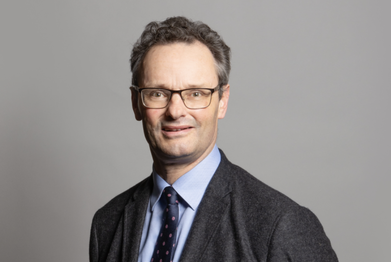 A man with curly hair and glasses smiles, wearing a dark blazer over a light blue shirt and a patterned tie against a gray background.