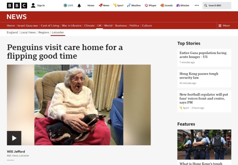 An elderly woman smiles and laughs while sitting in a chair, holding a black and white penguin on her lap. The scene is warm and joyful, highlighting a visit to a care home.