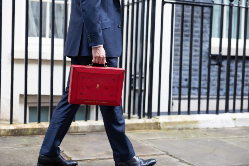 A man in a navy suit walks confidently down a street, carrying a red briefcase with gold accents.