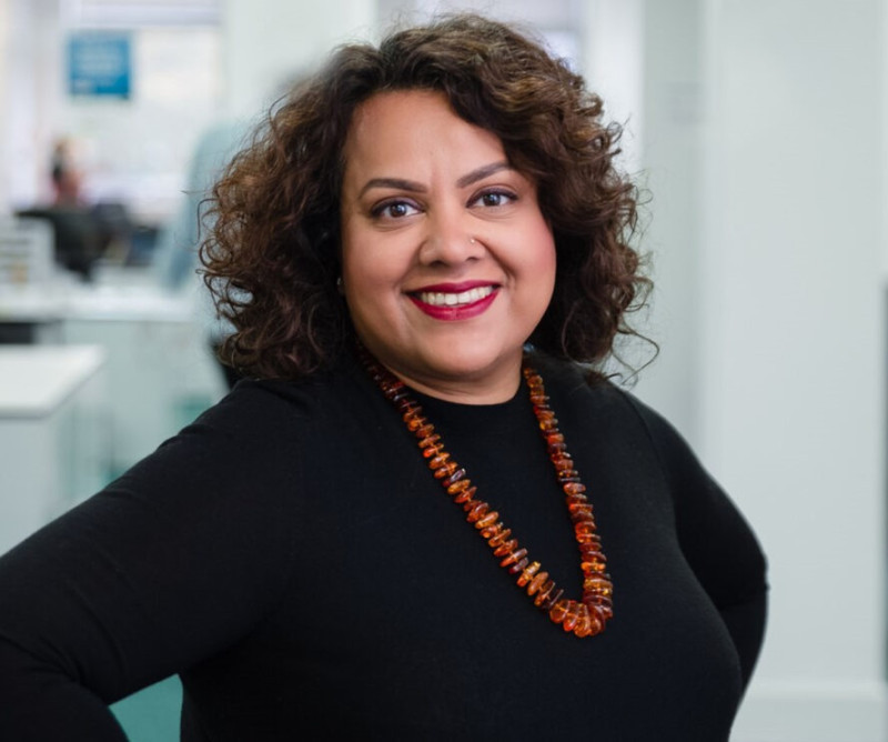 A smiling woman with curly hair poses confidently, wearing a black shirt and a long amber necklace. She stands in a bright, modern office environment.