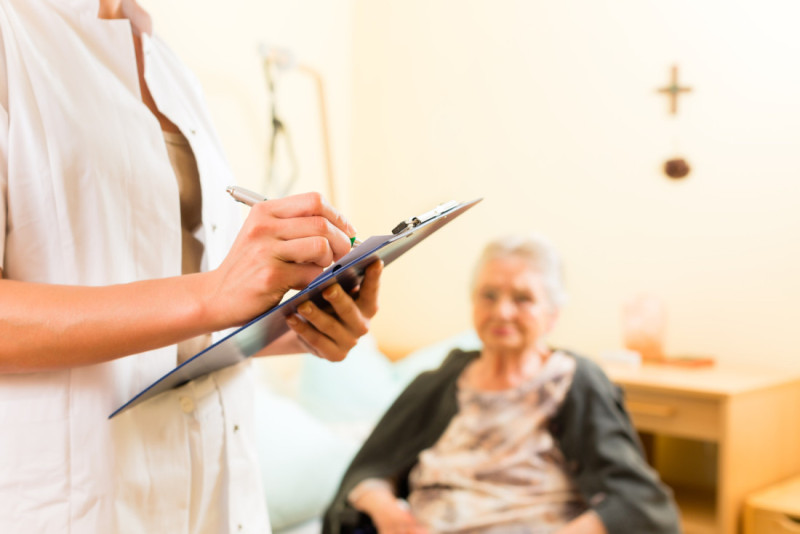 A healthcare professional writes notes on a clipboard while an elderly woman sits in a chair in the background, looking at her. Soft lighting creates a warm atmosphere in the room.