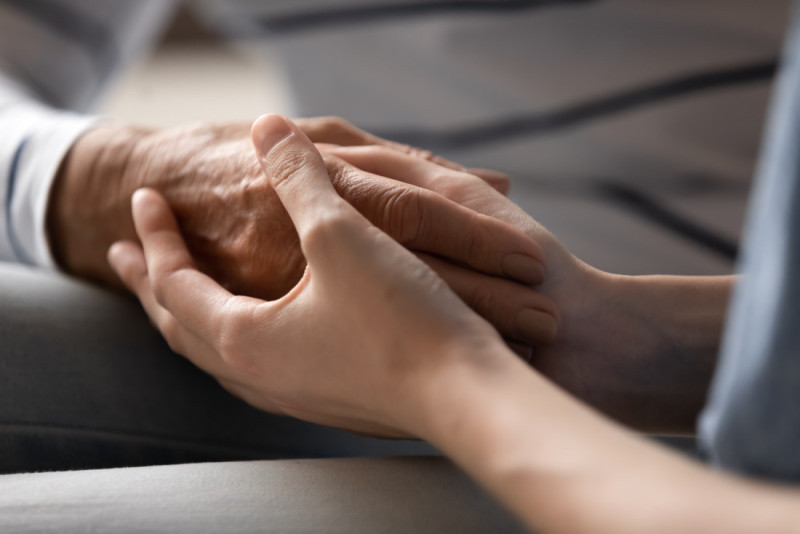 Two hands gently clasped together, conveying a sense of comfort and connection. One hand is older with visible wrinkles, while the other is younger and smoother.