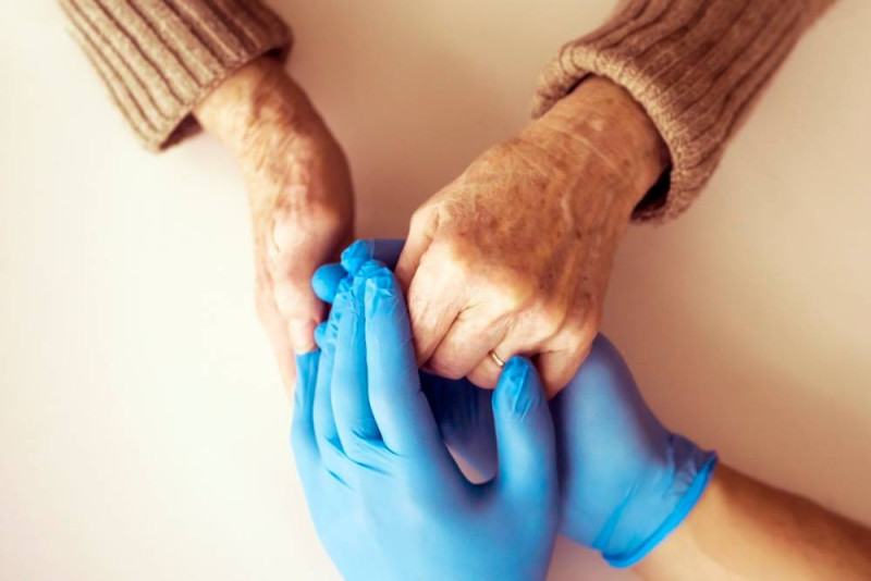 Two hands clasping each other, one is an elderly hand with visible wrinkles, and the other is a gloved hand, possibly a caregiver’s.