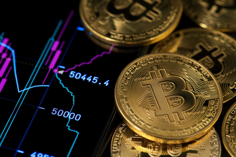 Bitcoin coins are stacked beside a digital graph displaying a price chart, with values around 50,445.4. The graph features purple and blue lines, indicating market trends.