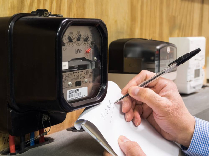 A person records energy meter readings in a notebook while observing a black electric meter.