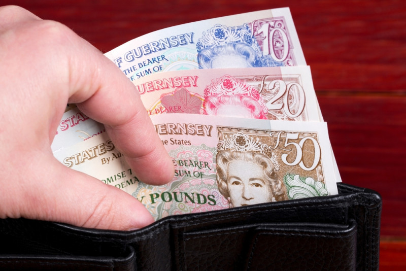 A hand reaching into a black wallet, pulling out several banknotes including £20, £50, and £70 notes from Guernsey.
