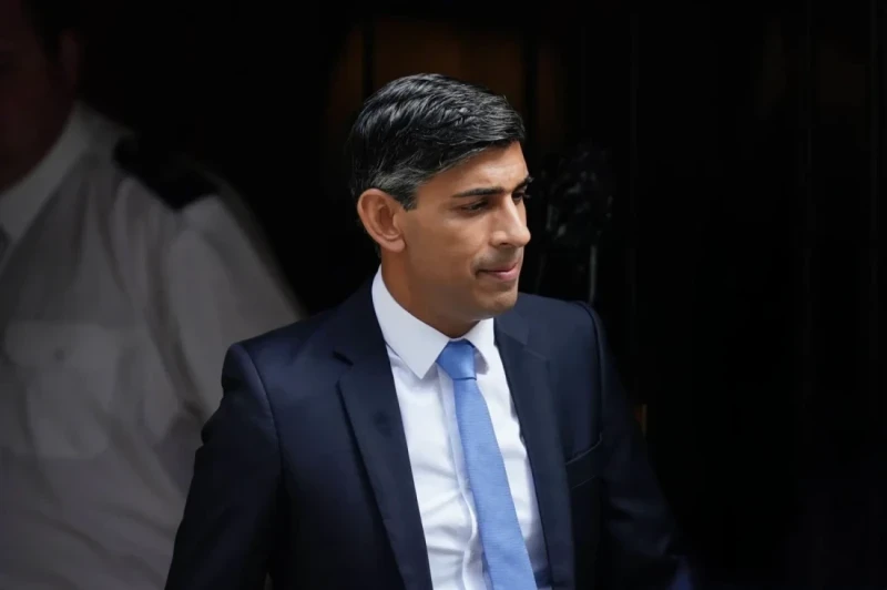 A man in a black suit and light blue tie is walking out of a building, looking slightly to the side. A uniformed officer is partially visible in the background.