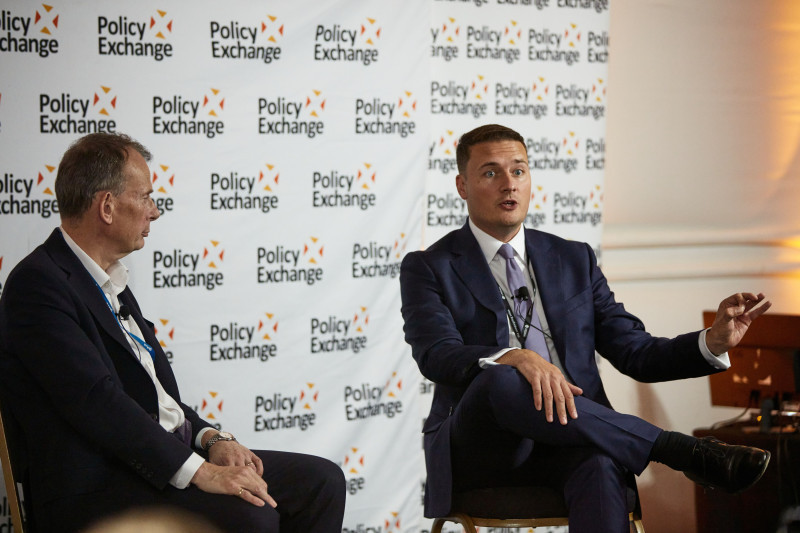 Two speakers engage in discussion during an event, seated in front of a backdrop featuring the Policy Exchange logo. One speaker gestures while making a point, and the other listens attentively.