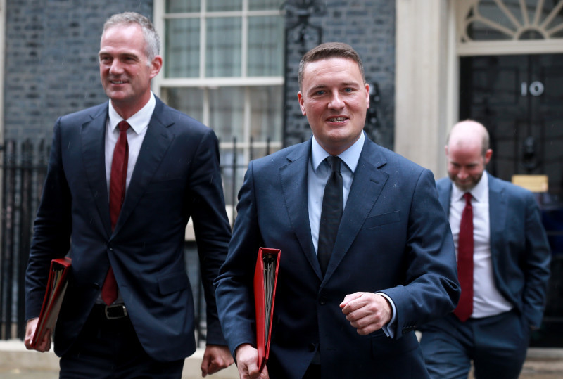 Three men in suits walk together outside a building with a black door. Two men are smiling and holding red folders, while one walks slightly behind them. The setting appears formal, with brick walls in the background.