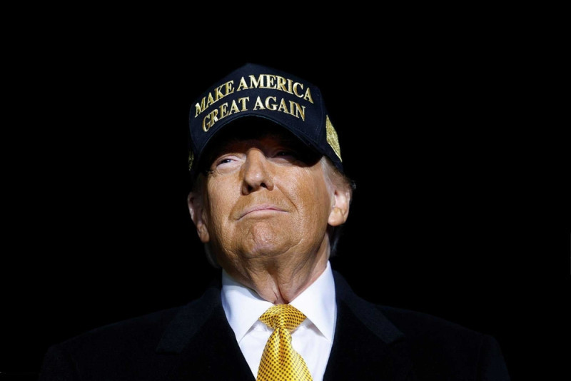 A man wearing a black cap with "Make America Great Again" written in gold, accompanied by a black suit and yellow tie, looking confidently ahead against a dark background.