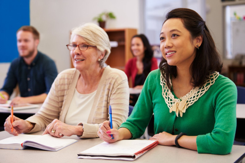 A diverse group of adults in a classroom setting, focused on their instructor. The foreground features a middle-aged woman with glasses and a senior woman taking notes. The background includes two other people listening attentively. A warm, collaborative atmosphere is evident.