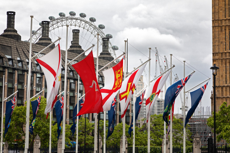 A row of flags, including the Union Jack and several regional flags, is displayed in front of buildings topped with chimneys. The London Eye and construction cranes are visible in the background under a cloudy sky.