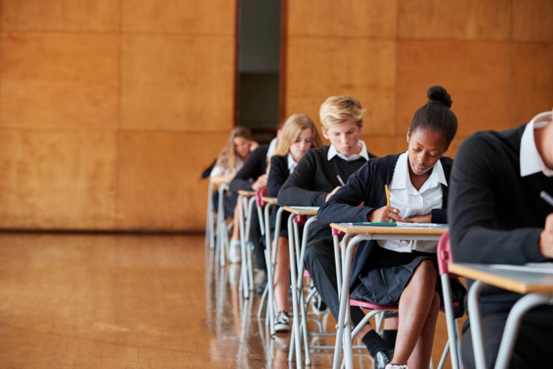 Students seated at desks in a classroom, focused on taking an exam. The room has wooden paneling and a polished floor.