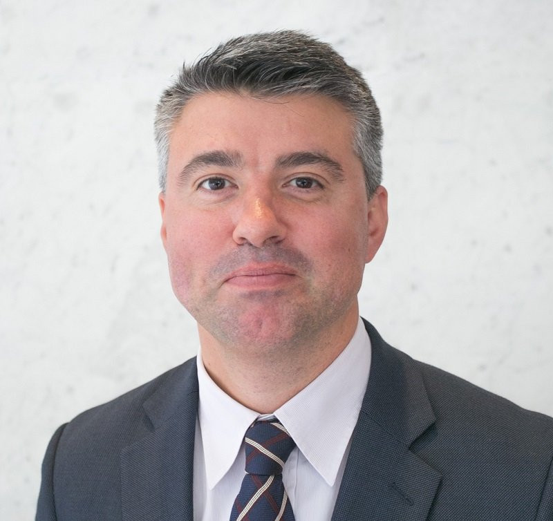 A man with short gray hair and a slight smile wears a dark suit and striped tie against a light textured background.