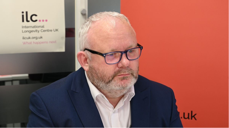 A man with a beard and glasses wearing a navy suit sits thoughtfully in front of a colorful backdrop featuring the logo of the International Longevity Centre UK.