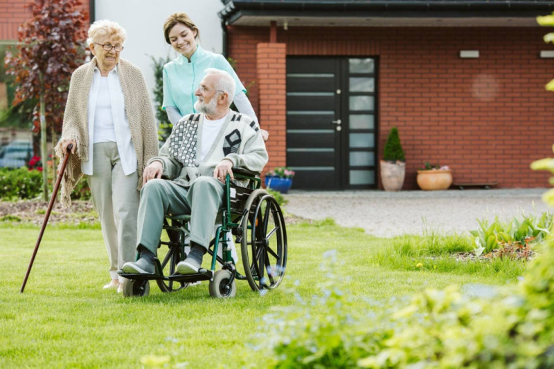 An elderly woman with glasses and a cane walks alongside a smiling caregiver, who pushes a man in a wheelchair through a garden with green grass and blooming plants.