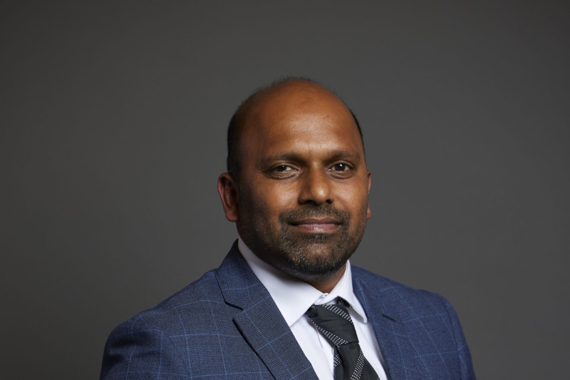 A man in a blue checked suit jacket and white shirt stands against a dark background, smiling confidently.