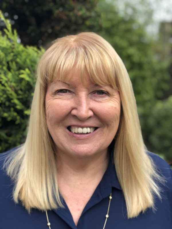 Smiling woman with long, straight blonde hair, wearing a navy blue shirt, standing outdoors with green foliage in the background.