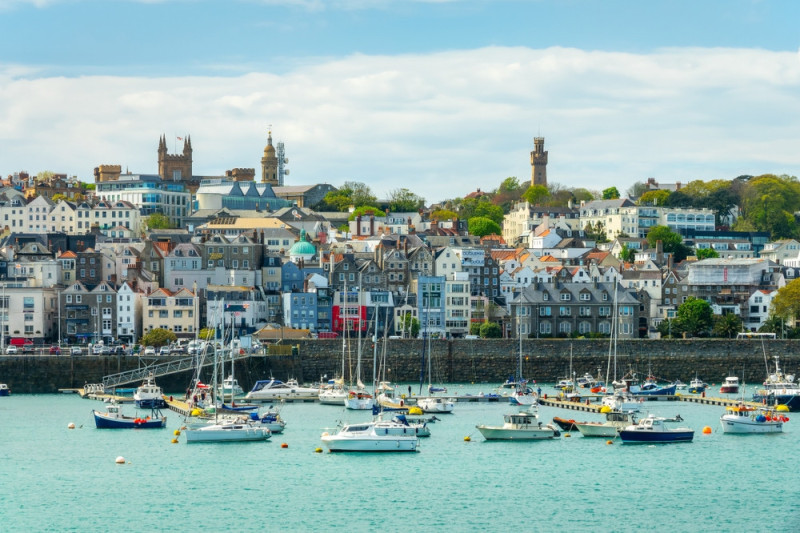 A scenic view of a coastal town with a bustling harbor filled with boats. Colorful buildings and lush greenery rise on the hillside, complemented by a blue sky and soft clouds. Notable landmarks can be seen among the structures, indicating a vibrant community.
