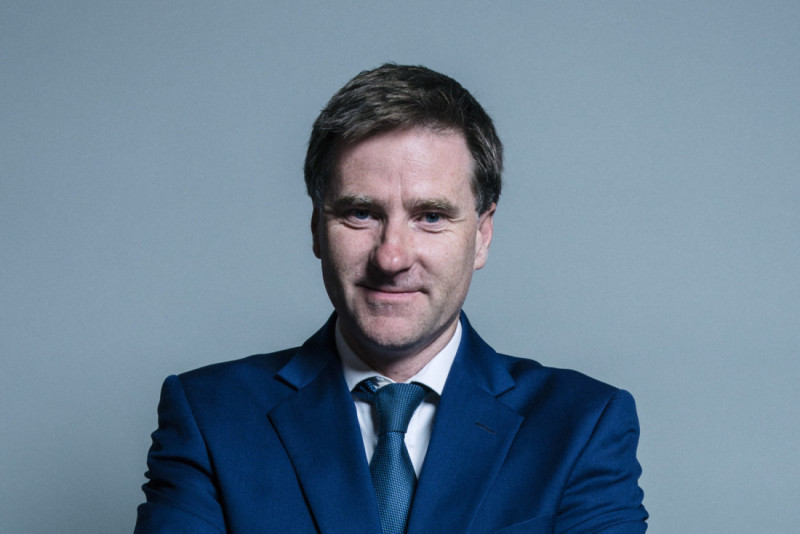 A man in a blue suit and tie stands confidently with his arms crossed, smiling slightly against a plain gray background.