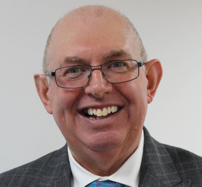 Smiling man with short gray hair and glasses, wearing a gray suit and a colorful tie, against a plain white background.