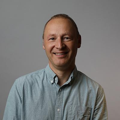 A smiling man with short, light brown hair wearing a button-up, light blue shirt against a gray background.