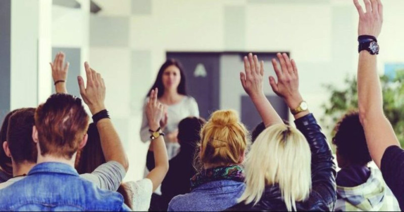 A group of people with raised hands, attentively listening to a speaker in a classroom or conference setting.