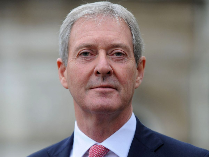 A middle-aged man in a suit and tie, with short gray hair, is looking directly at the camera with a neutral expression. The background is slightly blurred, suggesting an outdoor setting.